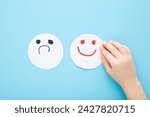 Baby hand with bad and happy smileys on light blue table background. Pastel color. Negative and positive smiling face expressions on white paper. Closeup. Children mood concept. Top view.