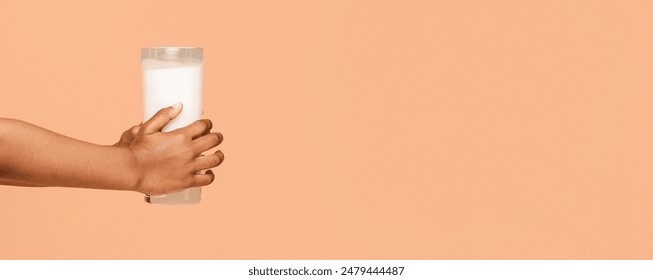 Baby hand is holding a glass of milk against a peach-colored background. The glass is filled with white milk, and the hand is shown from the wrist to the fingertips, panorama with copy space - Powered by Shutterstock