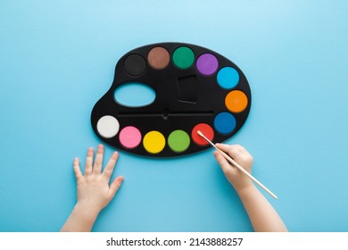 Baby Hand Holding Paint Brush And Using Colorful Palette On Light Blue Table Background. Pastel Color. Closeup. Point Of View Shot. 