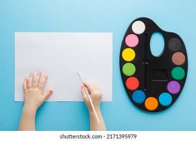 Baby Hand Holding Paint Brush. Colorful Palette And Empty White Paper On Light Blue Table Background. Pastel Color. Closeup. Point Of View Shot.