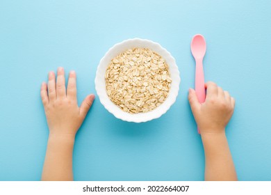 Baby Hand Holding Pink Plastic Spoon. White Bowl With Dry Rolled Oat On Light Blue Table Background. Pastel Color. Closeup. Point Of View Shot. Children Healthy Food. 