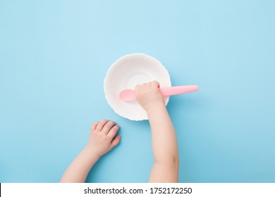 Baby hand putting pink plastic spoon in empty bowl and waiting food on light blue table background. Pastel color. Closeup. Point of view shot.