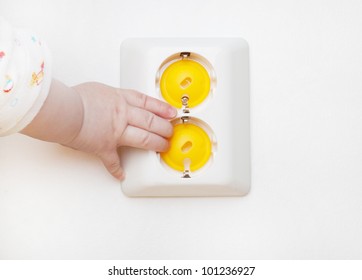 Baby Hand Reaching For An Electrical Outlet Covered With Yellow Safety Plugs (baby And Child Safety Concept)