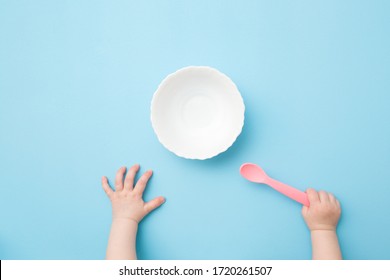 Baby Hands Holding Pink Plastic Spoon And Waiting Food. Empty White Bowl On Light Blue Table Background. Pastel Color. Closeup. Point Of View Shot.