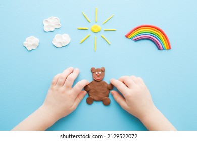 Baby hands making brown bear from modeling clay. Colorful rainbow, yellow sun and white clouds on light blue table background. Pastel color. Closeup. Point of view shot. Toddler development.