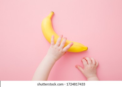 Baby Hands Taking Yellow Banana. Fresh Fruit On Light Pink Table Background. Pastel Color. Closeup. Point Of View Shot.