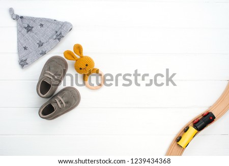 Baby hat, booties and wooden teether and train toy on white wooden background with blank space for text. Top view, flat lay.