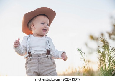 Baby In A Hat And Suspenders Looking In The Field At Sunset