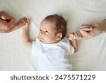 Baby holding parents hands. Father and mother hold newborn kid cute infant child in white bodysuit lying on white sheet on bed