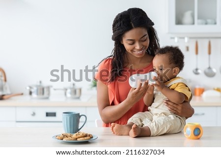 Baby Hydration Concept. Loving Black Mother Giving Water Bottle To Her Infant Child That Sitting On Kitchen Counter, Adorable Toddler Boy Enjoying Healthy Drink, Spending Time With Mom At Home