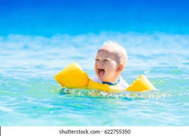 Baby With Inflatable Armbands Swimming In Ocean Water On Exotic Beach. Little Boy Wearing Sun Protection Rash Guard. Child With Swim Wings Floating In Tropical Sea. Family And Kids Summer Vacation.