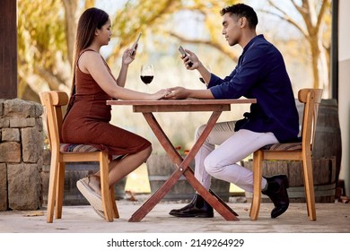 Baby, Just A Sec. I Have To Update My Socials. Shot Of A Young Couple Having Wine On A Date On A Wine Farm.