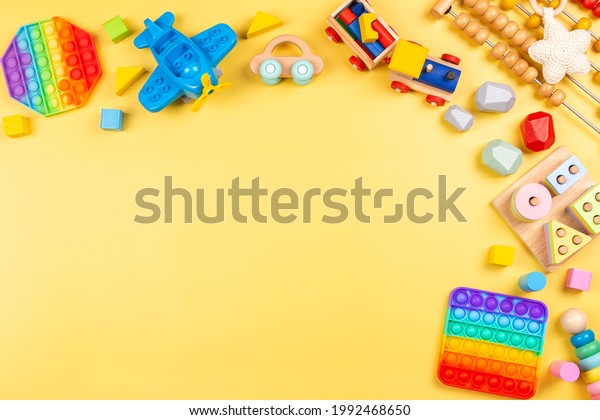Baby kids toys background with wooden toys, abacus,\
plane, pop it fidget toys and colorful blocks on yellow background.\
Top view, flat lay