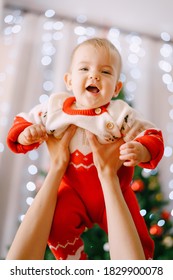 Baby In A Knitted Christmas Onesie In Her Mother's Arms In Front Of A Christmas Tree