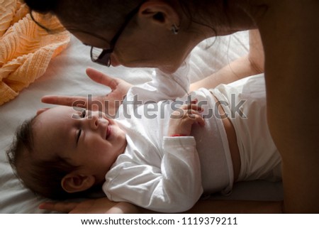Baby laying on the bed smiling at his mom who is leaning over him.