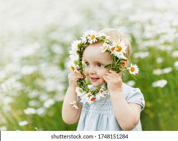 a baby in a light blue dress looks away through a wreath of cammomile. Stands in a chamomile field