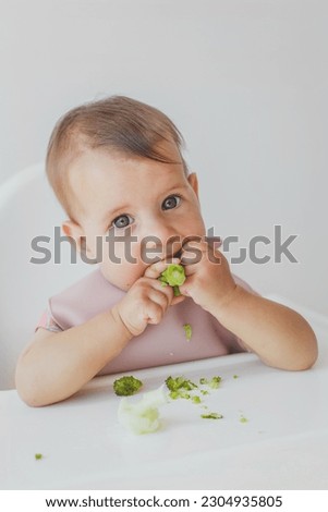 baby little girl 8 months old sits in a high chair and eats complementary foods green broccoli, close-up portrait looks at the camera. baby food concept. vertical photo