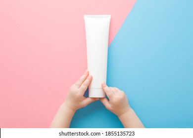 Baby Little Hands Holding White Cream Tube On Light Pink Blue Table Background. Pastel Color. Care About Clean And Soft Body Skin. Daily Children Beauty Product. Closeup. Point Of View Shot.