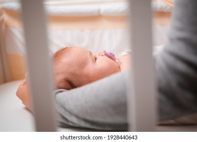 A Baby Is Lying In The Cradle With A Pacifier, The Mother Is Stroking His Head. Side View, Through The Bars Of The Cradle.