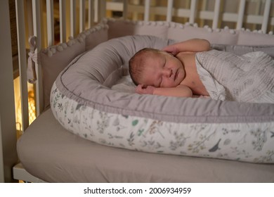 A Baby Newborn Is Sleeping In A Baby Cocoon With A Burning Night Light. A Child Is An Infant In A Home Crib By The Light Of A Flashlight