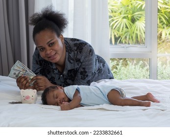 Baby newborn son sleeping on a white bed next to African mother who's putting money in a piggy bank. Saving money for baby's future. - Shutterstock ID 2233832661
