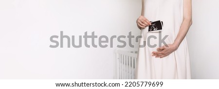 In baby nursery pregnant woman in white dress holding ultrasound image, baby sonography. Concept of pregnancy, health care, gynecology, medicine. Copy space. Happy lady enjoying photo of unborn child.