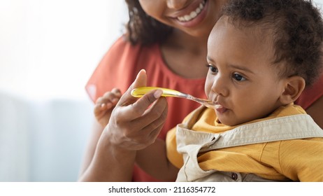 Baby Nutrition Habits. Smiling Black Mother Feeding Cute Infant Boy From Spoon, Young Caring African American Mom Giving Healthy Food To Her Adorable Toddler Child At Home, Closeup With Copy Space