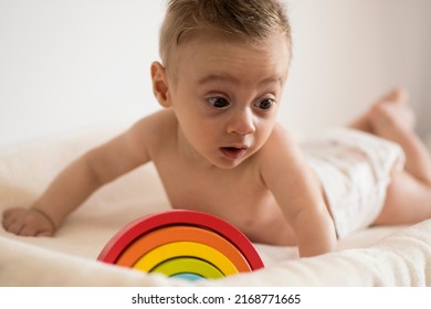 Baby On A White Bed Making Funny Faces To A Toy That Is A Rainbow