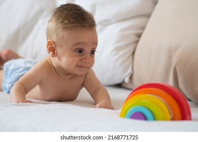 Baby On A White Bed Making Funny Faces To A Toy That Is A Rainbow