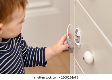 Baby Opens The Child Lock On The Closed Drawer Of The Cabinet. Toddler Baby With Hand Opens Door Lock On Chest Of Drawers. Kid Aged One Year And Three Months