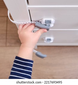 Baby Opens The Child Lock On The Closed Drawer Of The Cabinet. Toddler Baby With Hand Opens Door Lock On Chest Of Drawers. Kid Aged One Year And Three Months