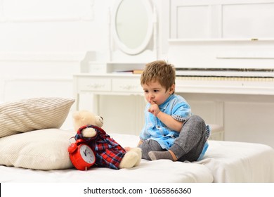 Baby Playing Bear Playing On White Bed. Kid Put Toy In Bed For Sleep, Shows Gesture To Be Silent. Teddy Bear Sleeps Near Alarm Clock.