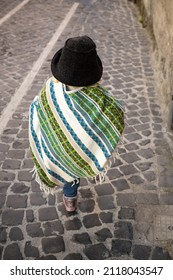 Baby In Poncho And Felt Hat Walking Ahead Along The Street With Cobblestone Pavement. View From Behind, No Face. Baby Fashion, Latin Stile. Colourful Striped Cape With Tassels. Independence Concept