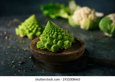 ein Babyromanesco-Broccoli auf einer Holzteller, auf einer dunklen Steinoberfläche neben einem anderen Babyromanesco-Broccoli-Kopf und einigen Blumenköpfen