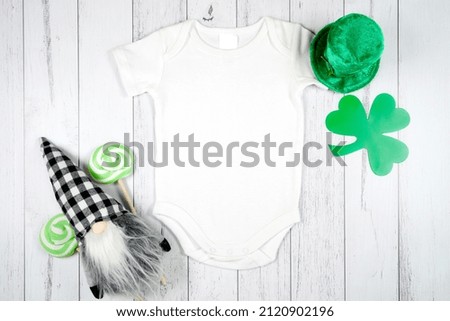 Baby romper onesie product mockup. St Patrick's Day farmhouse theme SVG craft product mockup styled with green leprechaun hat and buffalo plaid gnome against a white wood background.