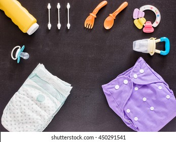 Baby set of cloth diaper, disposable diaper, baby powder, tither, cotton buds, spoons, soother and nibbler on dark background with copy space. Top view or flat lay