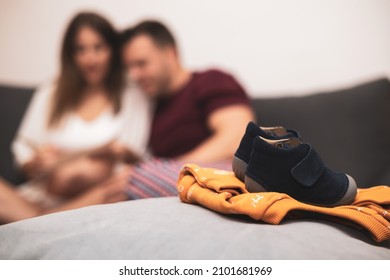a baby shoe.in the background a wedding couple sitting on the couch at home.the woman is pregnant.