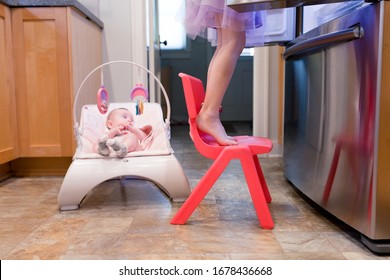 A Baby Is Sitting In A Bouncy Seat Watching Her Older Sister Make Lunch.