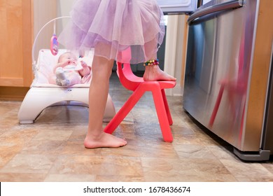 A Baby Is Sitting In A Bouncy Seat Watching Her Older Sister Make Lunch.