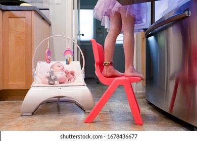 A Baby Is Sitting In A Bouncy Seat Watching Her Older Sister Make Lunch.