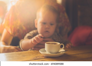 A Baby Is Sitting At A Table In A Cafe And Is Reaching For His Mother's Cup Of Coffee