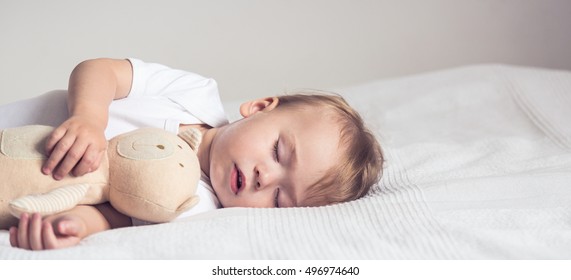 Baby sleeping in crib. Peaceful baby lying on a bed while sleeping in a bright room with his teddy bear. Panoramic view.