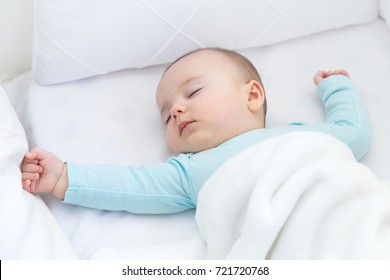 Baby Sleeping With Open Arms In A Cradle. Light Blue Pajama And White Bed Sheets.