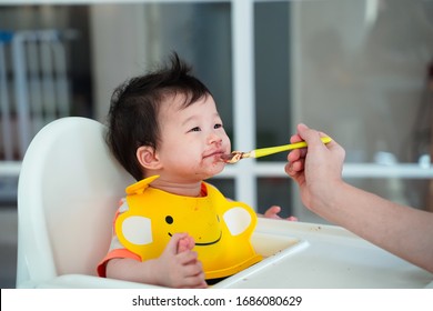 Baby Smiling While Eating With Mother Hand Holding Small Spoon Feeding Baby,toddler's Messy Wearing Yellow Bib Apron And Sitting In High Chair,solid Food For Little Baby.parent Feed Healthy Nutrition.