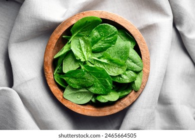 Baby spinach leaves with water drops in a wooden bowl on linen tablecloth. Raw organic spinach greens close up