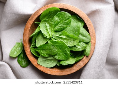 Baby spinach leaves with water drops in a wooden bowl on linen tablecloth. Raw organic spinach greens close up