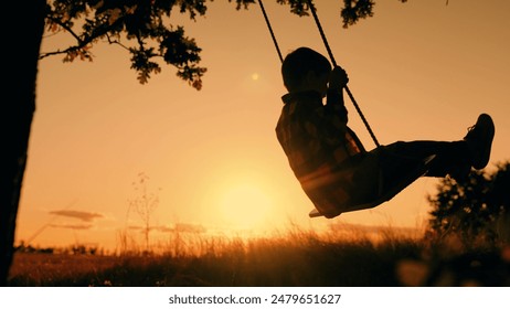 Baby swing, kid boy in flight. Child plays on wooden swing, dreams of fly. Happy little boy swings on swing in park under tree at sunset. Concept of family happiness, dreams, entertainment. Silhouette - Powered by Shutterstock