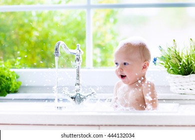 Baby Taking Bath In Kitchen Sink. Child Playing With Foam And Soap Bubbles In Sunny Bathroom With Window. Little Boy Bathing. Water Fun For Kids. Hygiene And Skin Care For Children. Bath Room Interior