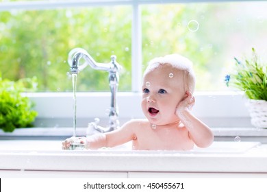 Baby Taking Bath In Kitchen Sink. Child Playing With Foam And Soap Bubbles In Sunny Bathroom With Window. Little Boy Bathing. Water Fun For Kids. Hygiene And Skin Care For Children. Bath Room Interior