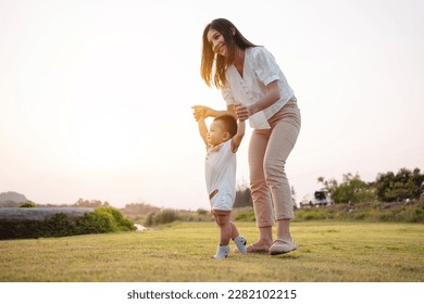 Baby taking first steps in park on meadow grass at sunset. Mother supports child to learn walking forward, baby growth and development concept. - Powered by Shutterstock